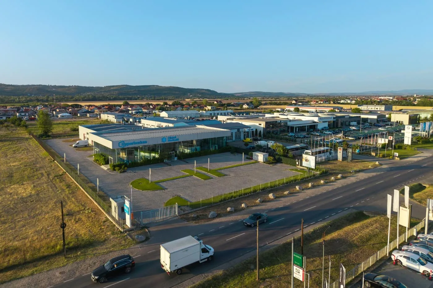 Wide aerial view of Oradea Medical Campus at golden hour, the paved car park and glass-fronted clinic beside the Calea Clujului frontage